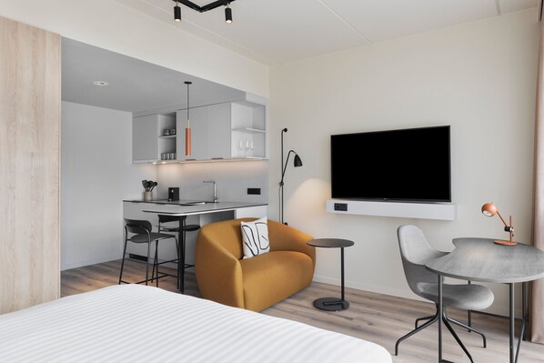 A modern hotel room with a bed, a sofa, a dining area with a table and chairs, a TV mounted on the wall, and a kitchen area.