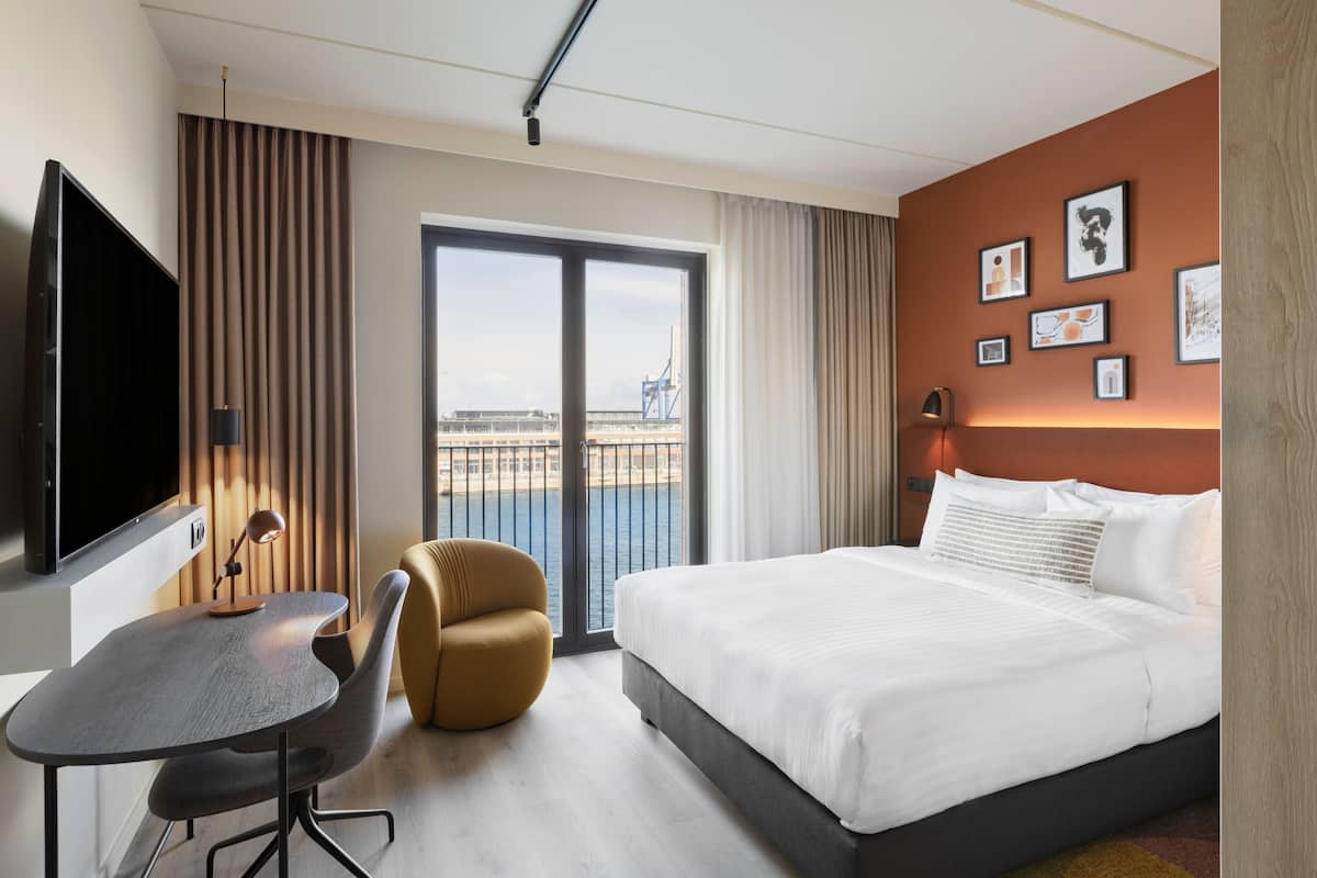 A hotel room with a bed, a desk, a chair, a TV, and a balcony with a view of the city.