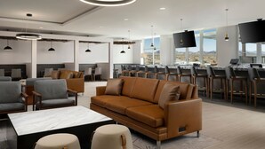 Un moderno vestíbulo de hotel con un sofá de cuero marrón, sillones grises, una mesa de centro de mármol y una zona de bar con taburetes y una gran pantalla de televisión.
