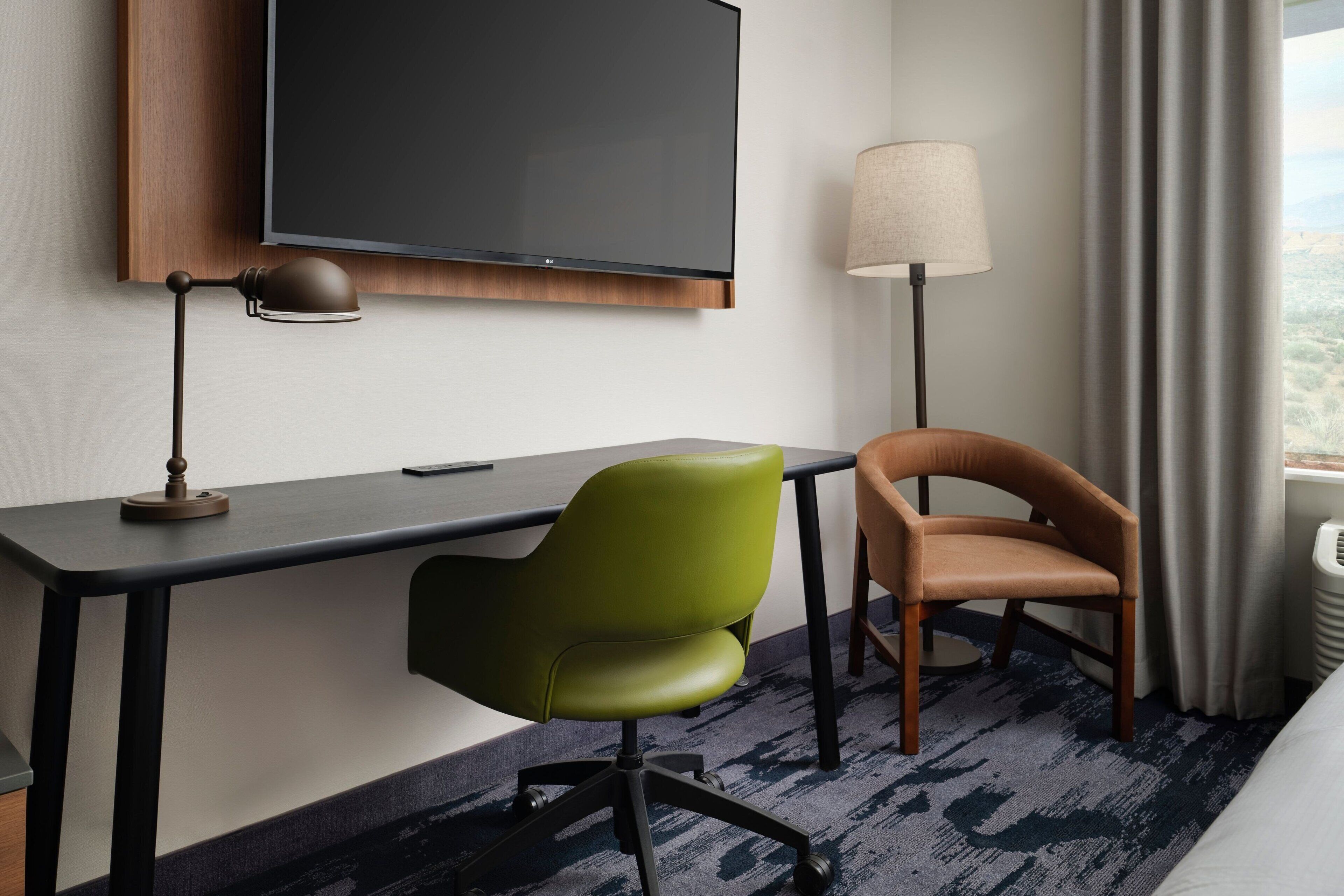 Quarto de hotel com televisão de tela plana na parede, escrivaninha com luminária, uma cadeira verde e uma marrom, e vista para o exterior através de uma janela com cortinas.