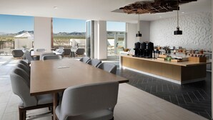 Un moderno comedor con una larga mesa y sillas, una encimera de cocina con varios electrodomésticos y grandes ventanas que ofrecen vistas del paisaje desértico.