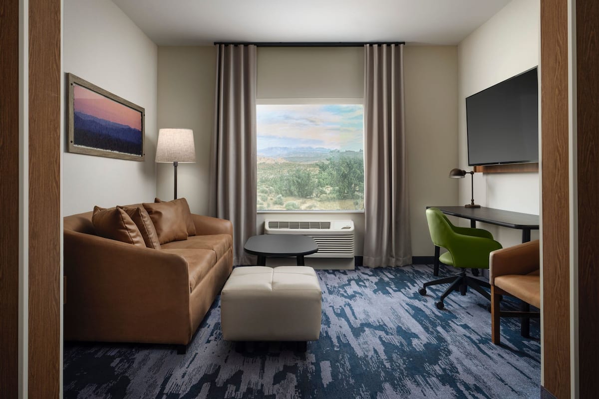 A hotel room with a brown leather sofa, a white ottoman, a green chair, a small table, a TV, and a large window with a view of the outdoors.