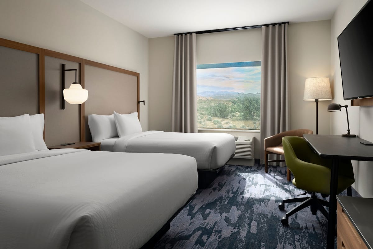 A hotel room with two beds, a desk, a chair, a TV, and a large window with a view.