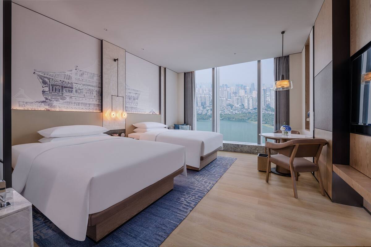A modern hotel room with two beds, a desk, and a large window offering a city view.