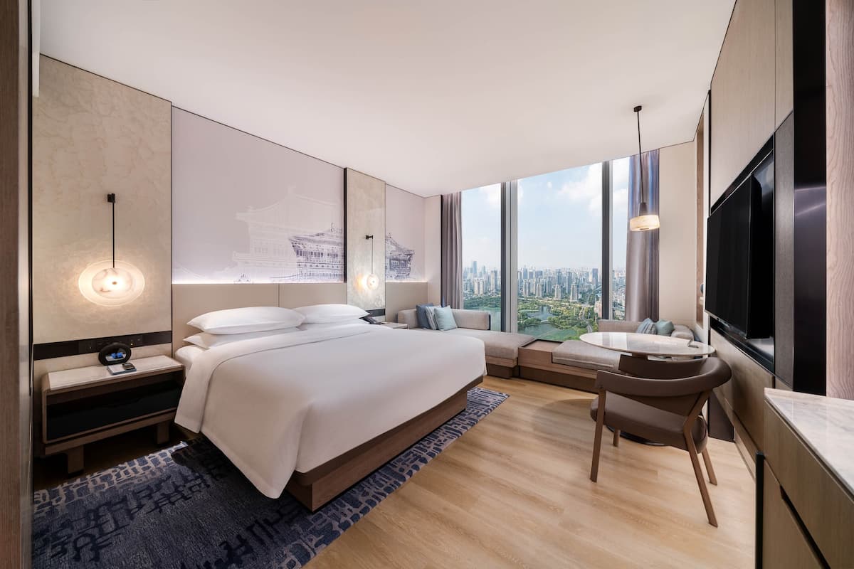 A modern hotel room with a large bed, a desk with a chair, a TV, and a city view.