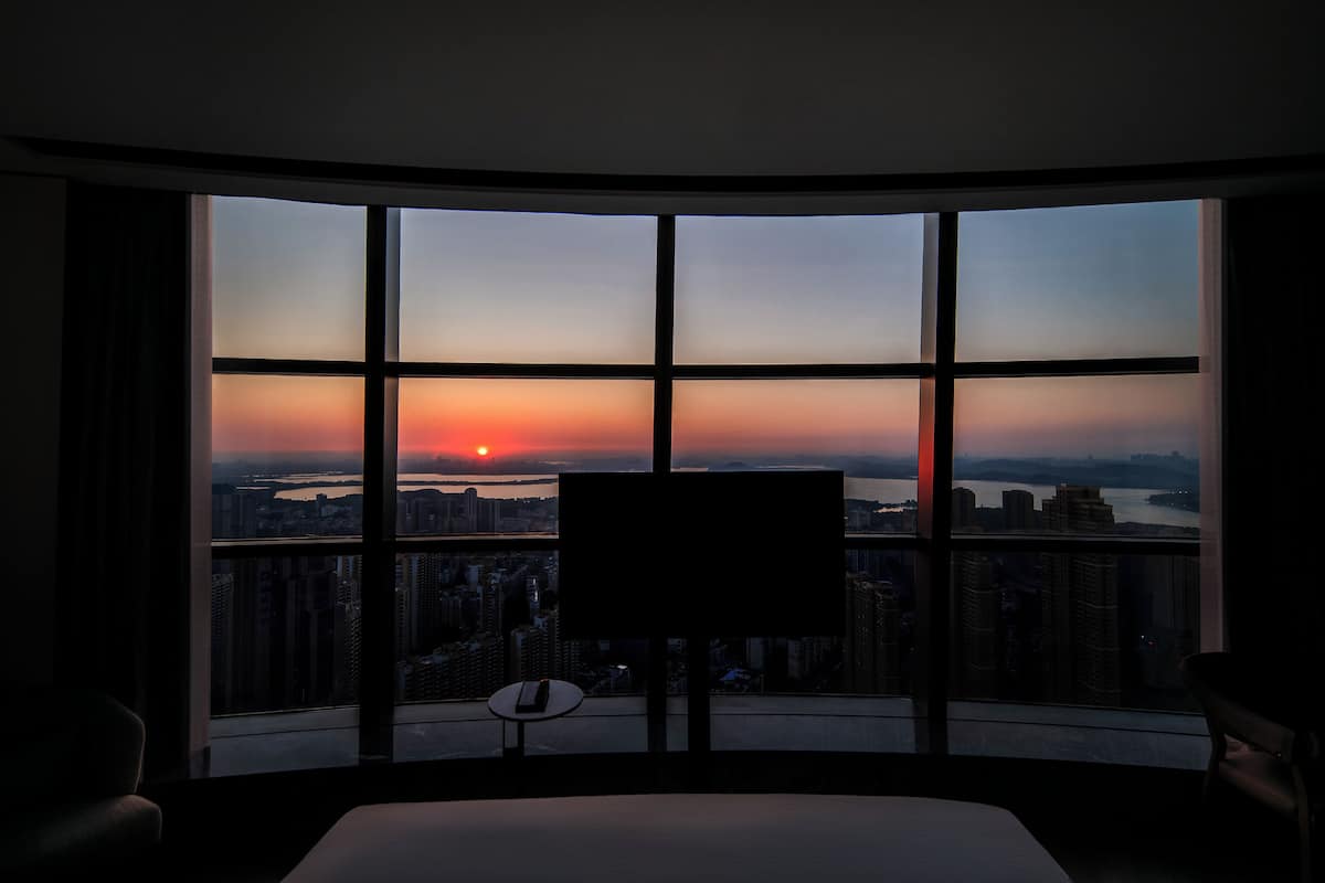 A hotel room with a large window offering a city view at sunset.