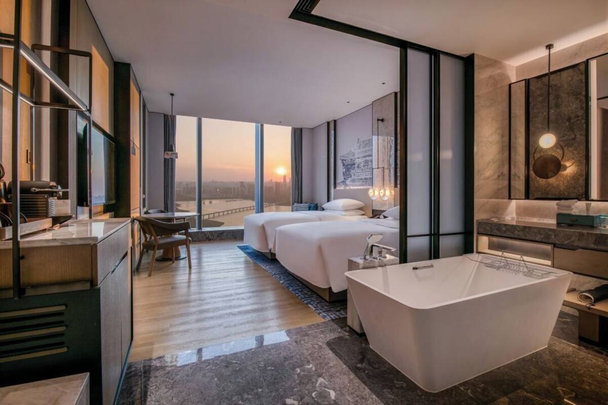 A modern hotel room with a large bathtub, two beds, a desk, and a view of the cityscape.