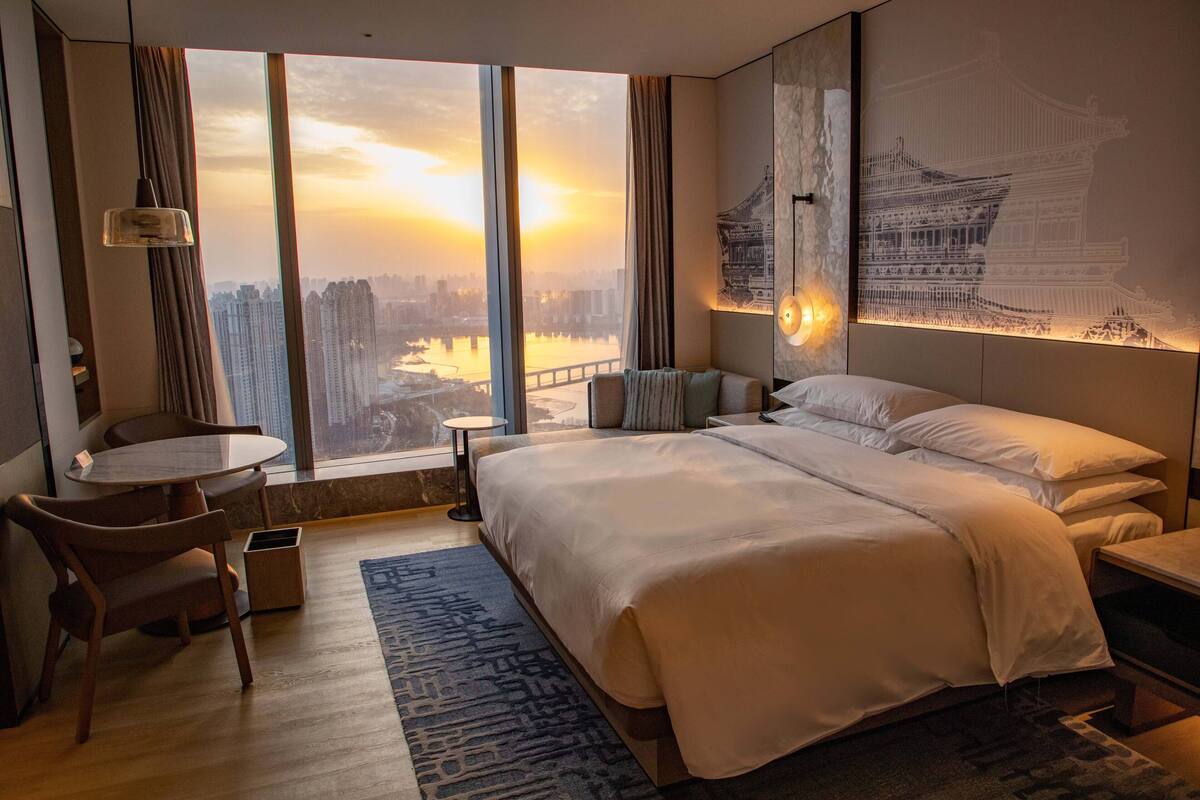 A hotel room with a large bed, a desk, and a view of a cityscape through large windows.