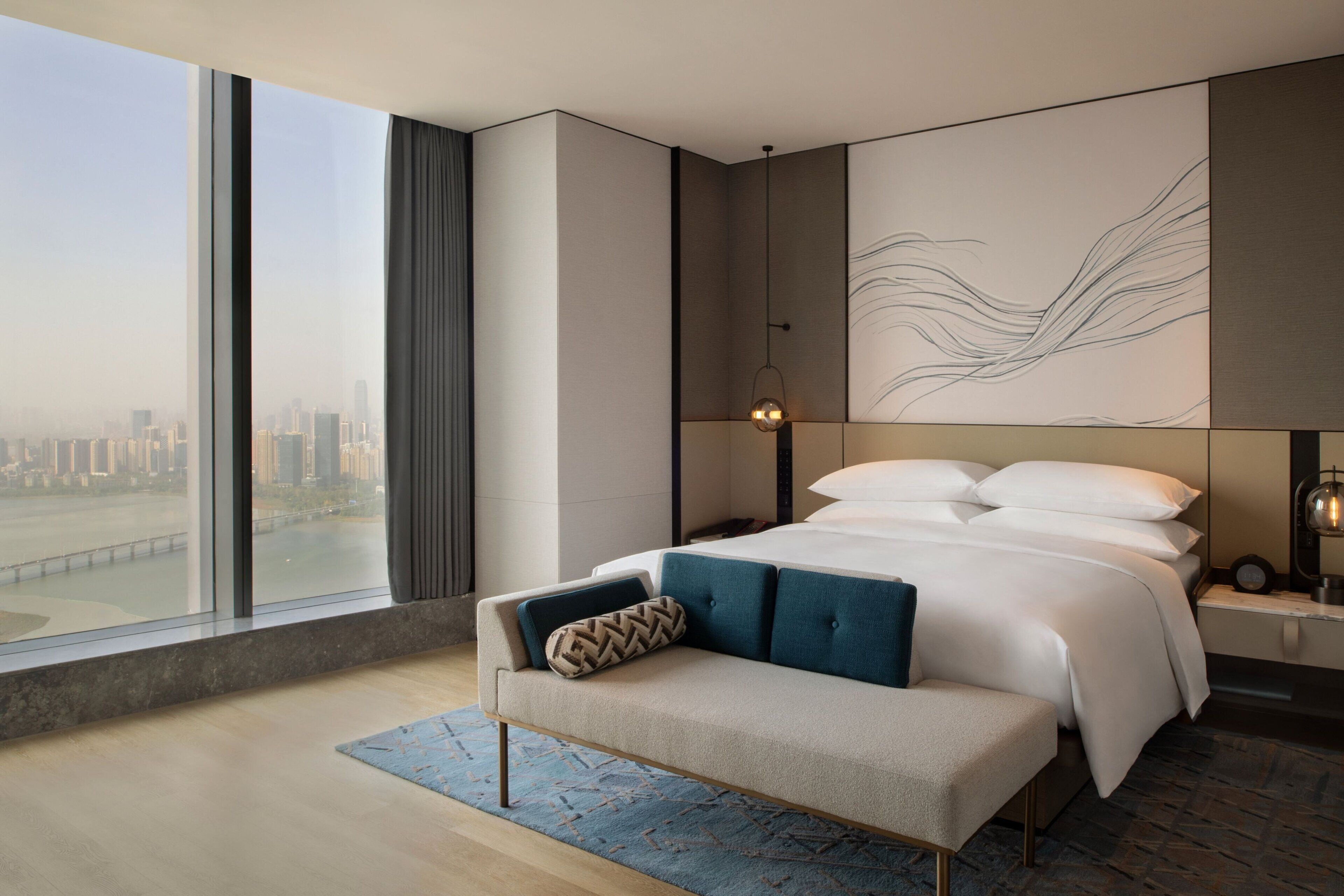 A modern bedroom with a large bed, a bench, and a city view.