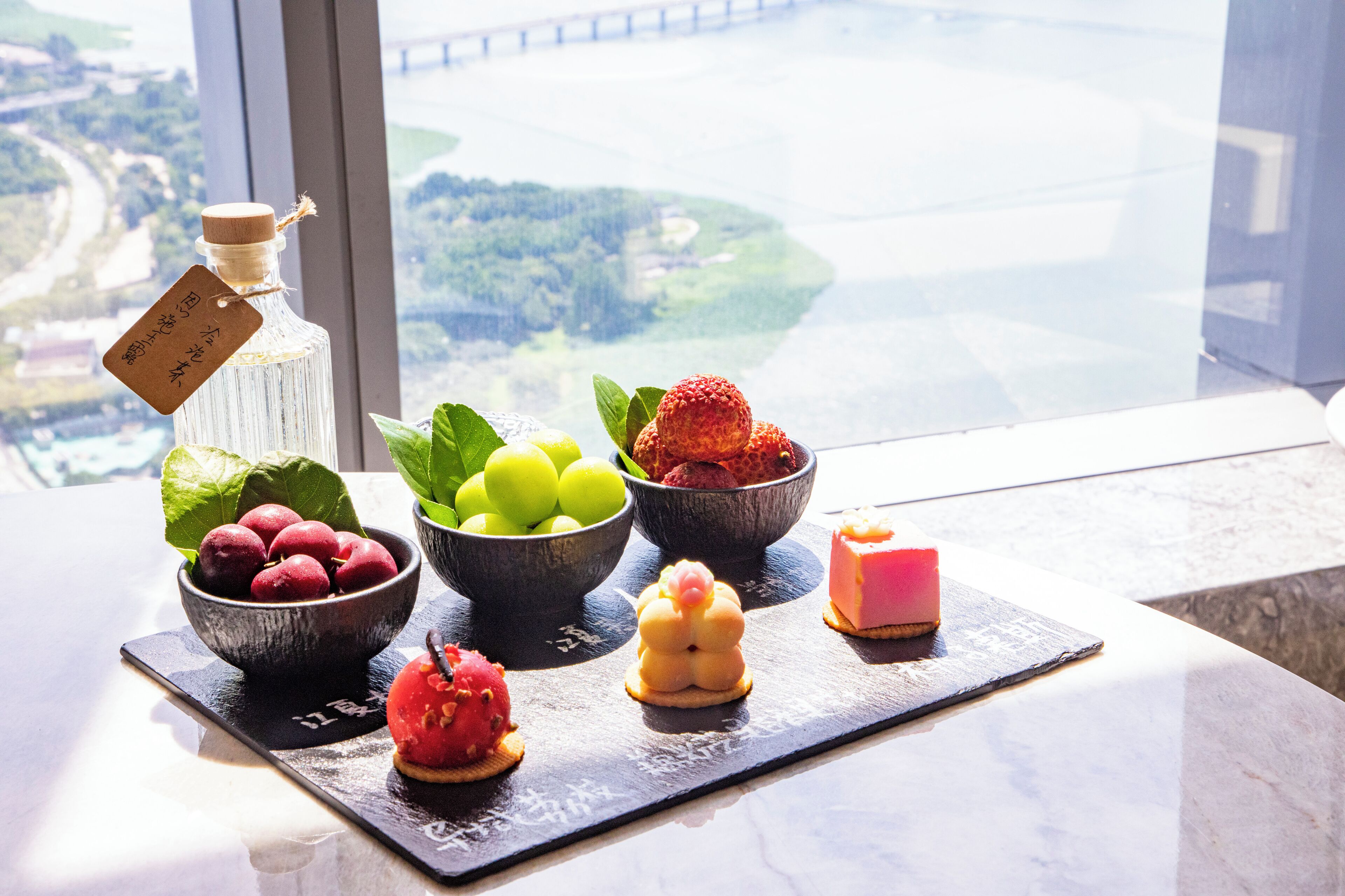 Un tavolo con frutta e dolci diversi, una bottiglia con un'etichetta, e vista su un ponte e del verde attraverso una finestra.