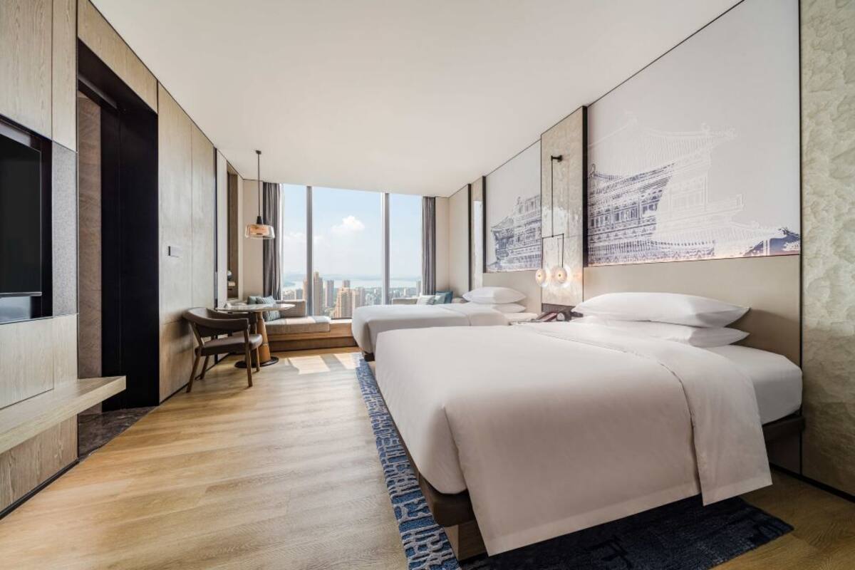 A modern hotel room with two beds, a dining area, and a large window offering a city view.