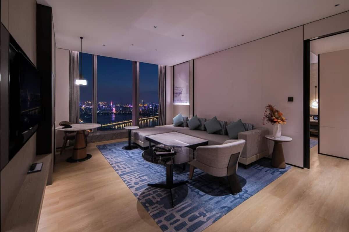 A modern living room with a sofa, coffee table, and TV, a city view through large windows, and a blue patterned rug.