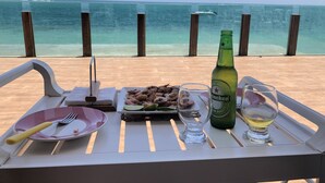 A dining table with a plate of seafood, a bottle of beer, and a glass of white wine, set against a beach view. - Village no Condominio Arauá, Fechado com Trilha Para Caminhada, Academia (Condomínio Arauá Vera Cruz)