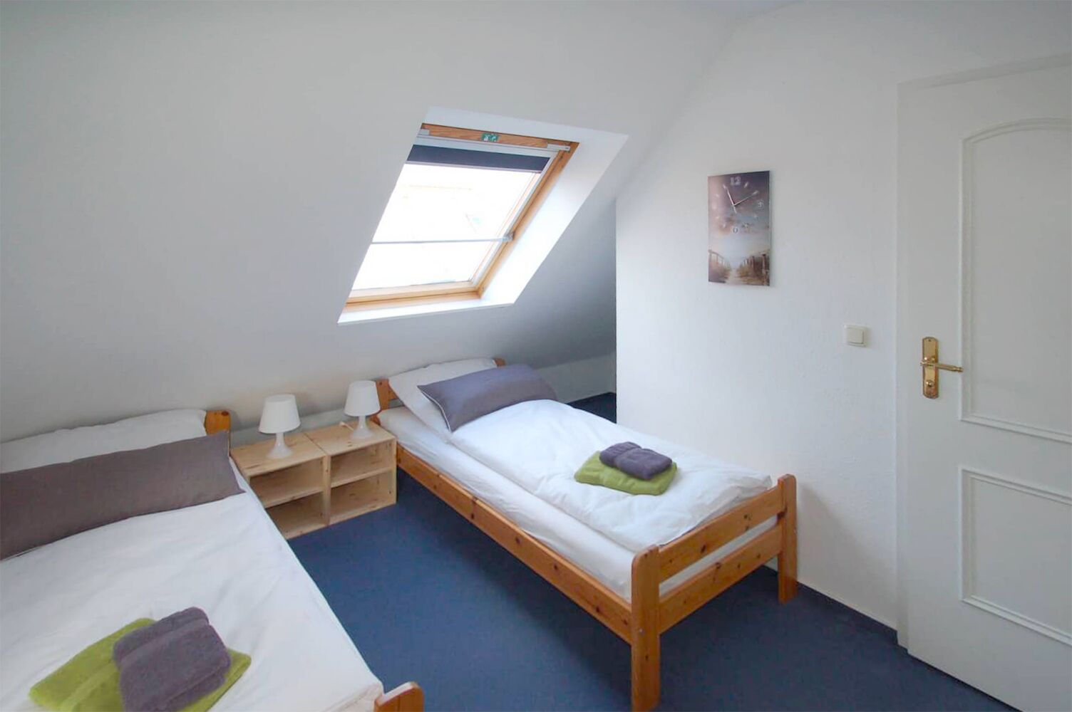A small, well-lit room with a sloped ceiling and a skylight, featuring two beds, a bedside table with lamps, and a door.