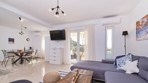 Un salon moderne comprenant un canapé, une table à manger avec des chaises, un téléviseur fixé au mur et un balcon offrant une vue sur l’extérieur.