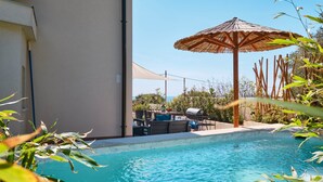 Une piscine agrémentée d’un parasol en bois, de chaises longues et d’un barbecue.