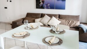 Uma área de refeições com uma mesa branca posta para quatro pessoas, um sofá marrom e uma pintura de paisagem urbana na parede.