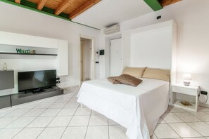 A bedroom with a bed, a TV, a bedside table, and a wall-mounted shelf with a 'Welcome' sign.