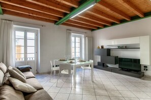 A modern living room with a dining table, white chairs, a sofa, a flat-screen TV, and a 'Welcome' sign.
