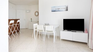 Ein modernes Wohnzimmer mit Flachbildfernseher, einem Essbereich mit weißen Stühlen und einer Küche im Hintergrund.