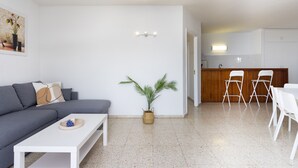 Ein modernes Wohnzimmer mit einem grauen Sofa, einem weißen Couchtisch und einer kleinen Pflanze. Es gibt eine Küchenzeile mit einer Holzarbeitsplatte und weißen Stühlen.