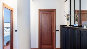 Un pasillo con una puerta de madera, un mueble oscuro con un espejo, y una cama visible a través de otra puerta abierta.
