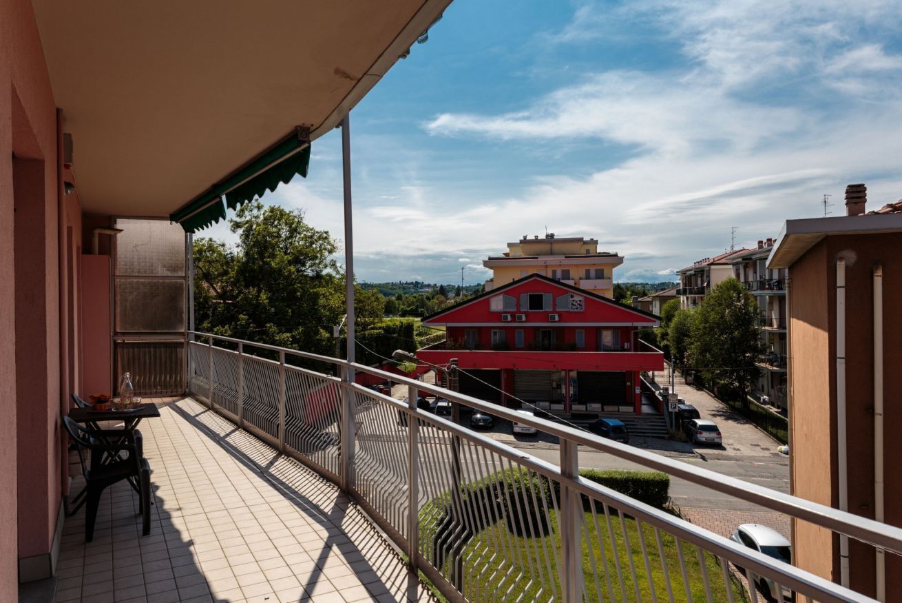 A balcony with a table and chairs, a view of a red building, and a street with parked cars.