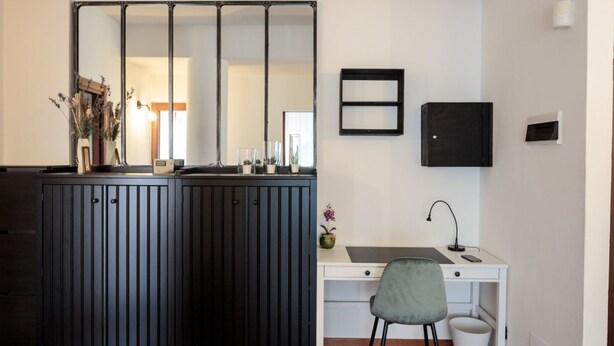 Una habitación compacta con un mueble de madera oscura, un escritorio blanco, una silla gris, un cubo de basura pequeño y un aplique de pared.