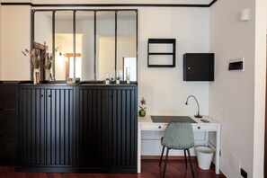 Een compacte kamer met een donker houten kastje, een wit bureau, een grijze stoel, een kleine prullenbak en een wandlamp.