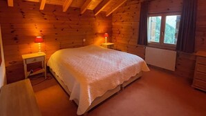 A wooden cabin bedroom with a bed, two bedside lamps, a window, and a radiator. - Large, very bright chalet for 8 people with large terrace, garden and a beautiful open view.Interior (St-Luc)