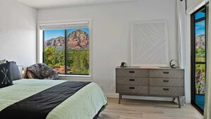 A bedroom with a bed, a dresser, a window with a mountain view, and a geometric wall art piece. - Spectacular Red Rock Views in West Sedona! (Sedona)