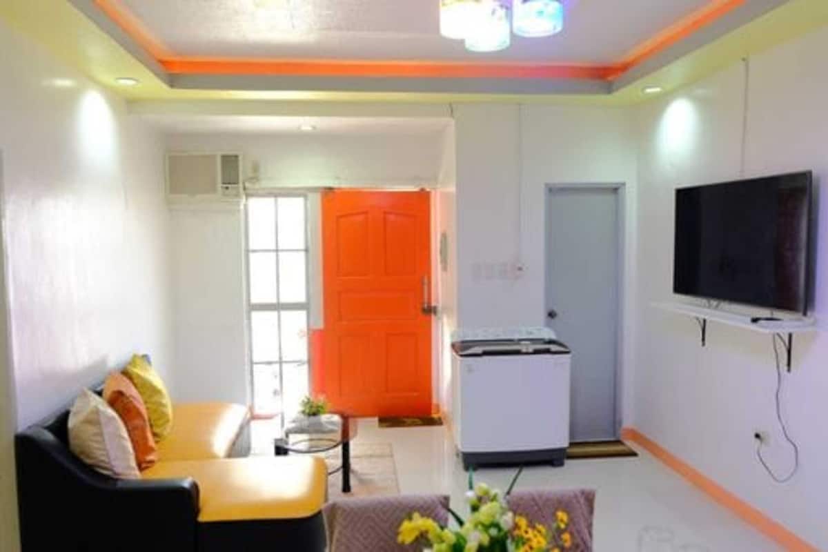 Ein kompakter Wohnraum mit einer Couch, einem kleinen Tisch, einem an der Wand befestigten Fernseher und einer leuchtend orangefarbenen Tür.