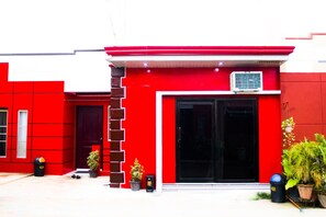 Un bâtiment rouge avec une porte et des fenêtres noires. On peut voir des plantes en pot et une poubelle à l’extérieur.