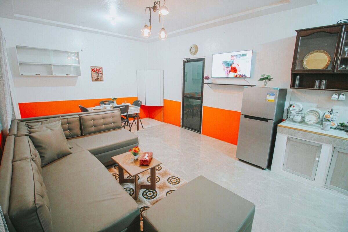 Ein modernes Wohnzimmer mit einer Couch, einem Couchtisch und einem Fernseher. Es gibt eine Küchenzeile mit einem Kühlschrank und einer Mikrowelle.