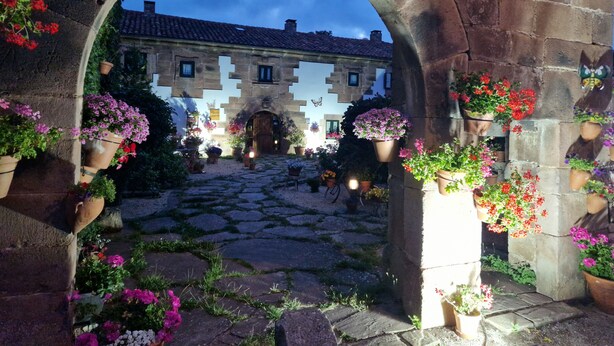 Un arco de entrada que da a un patio con pavimento de piedra, flores en macetas y un edificio con tejado de baldosas al fondo.