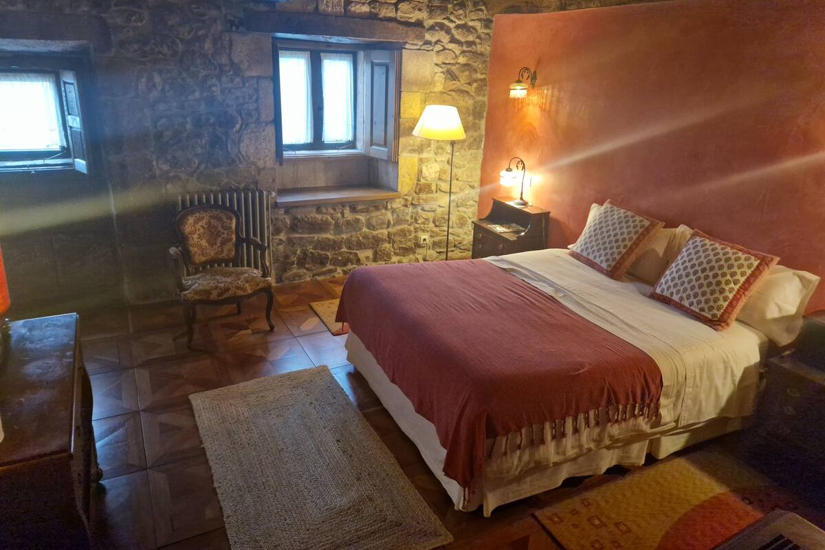 Une chambre confortable avec un mur en pierre, une chaise en bois, un lit agrémenté d’oreillers à motifs et une table de chevet avec une lampe.