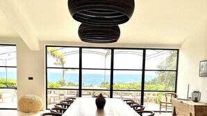 A long wooden table with chairs, a hanging light fixture, and large windows with a view of greenery and mountains. - Luxury villa in Gordes With Exceptional Views, Not Overlooked, Sleeps 16 (MURS)