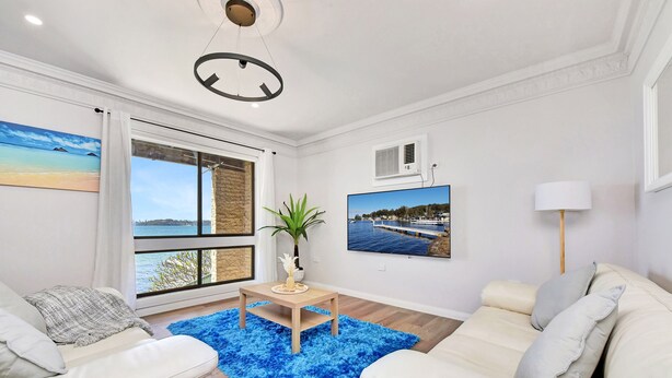 A modern living room with a white sofa, a blue rug, and a television displaying a coastal scene. - Silverwater Serenity Shores Absolute Waterfront (Silverwater)