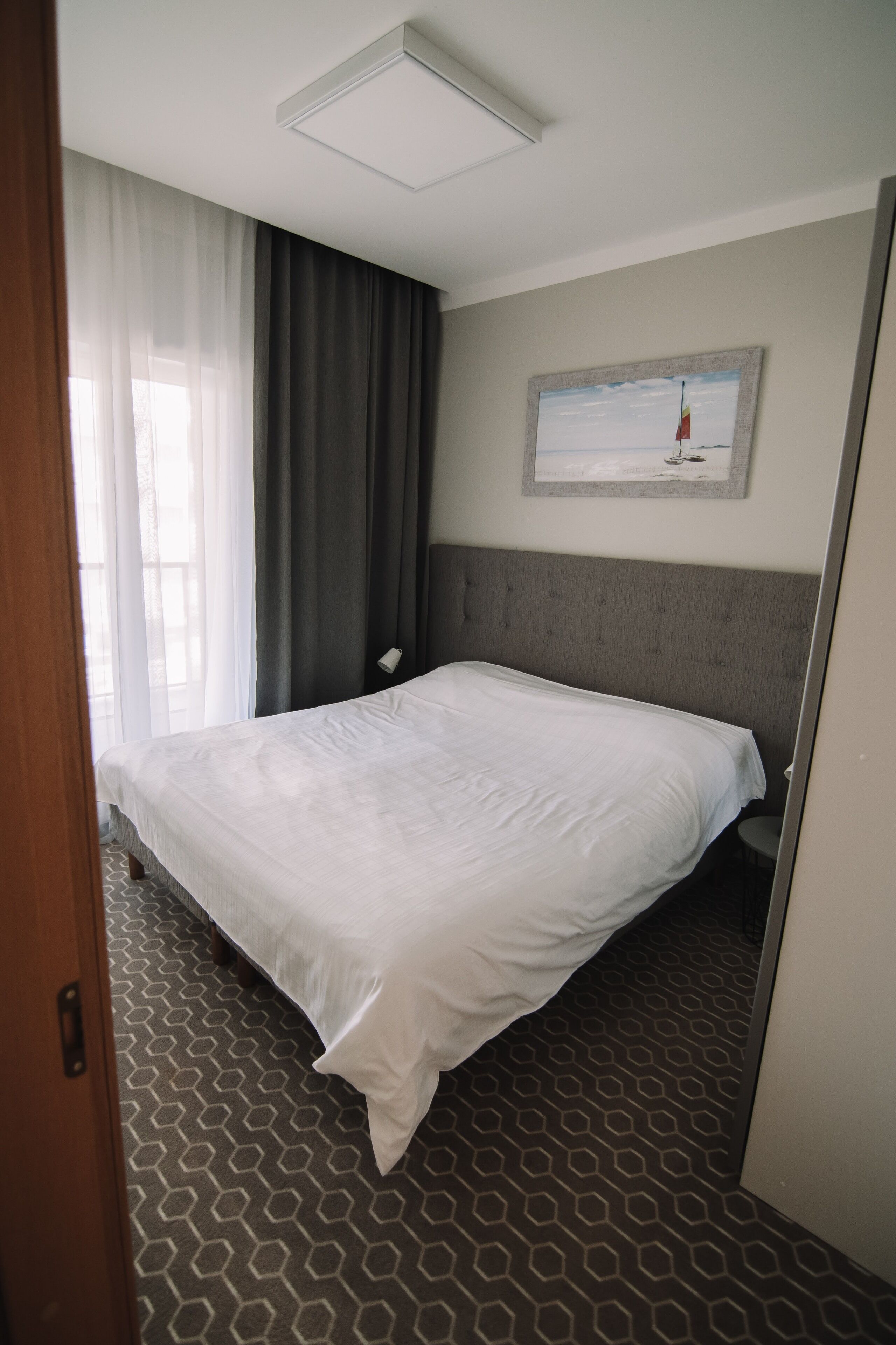 Una habitación de hotel con una cama grande, un cabecero, un cuadro en la pared y una ventana con cortinas.
