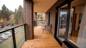 Une terrasse en bois avec une porte vitrée donnant sur une pièce, une petite table et une chaise, et une vue sur les voitures garées et les arbres.