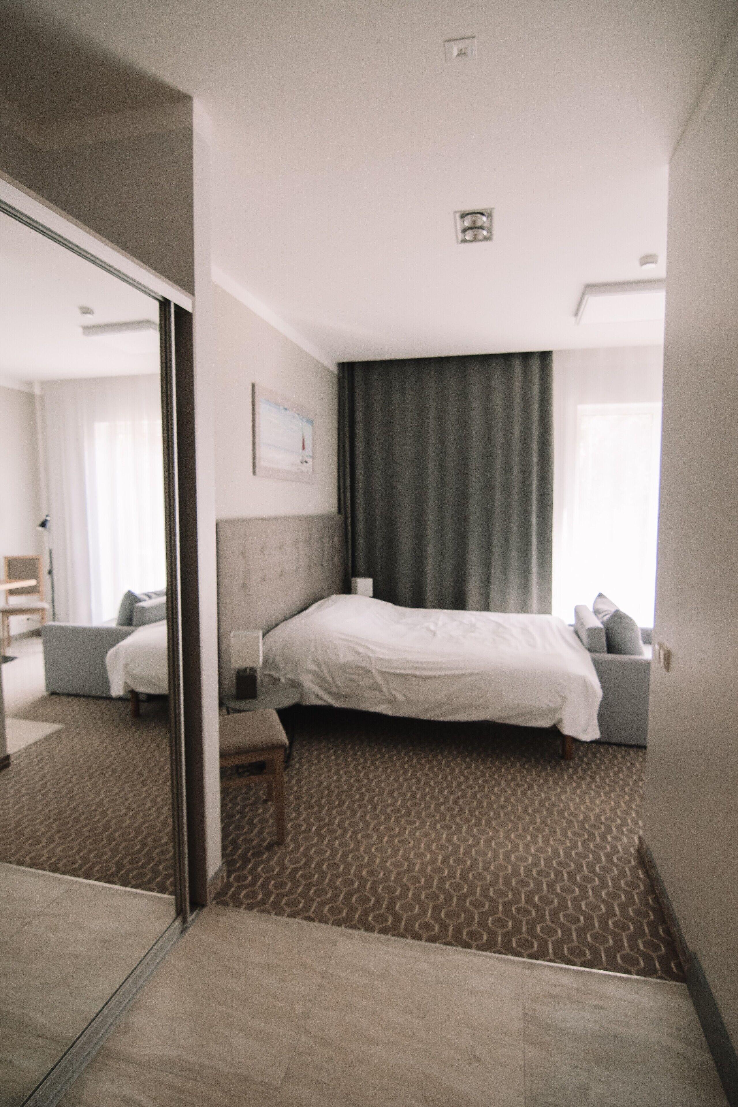 A modern hotel room with a large bed, a bedside table, a chair, and a mirror.