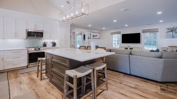 A modern kitchen with white cabinets, a central island with stools, a built-in oven, and a large island with a flat-screen TV. - 12 Heron Street- The Happy Heron (Hilton Head Island)