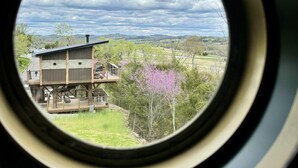 A view through a circular window of a house with a deck, surrounded by trees and a cloudy sky. - Two Treehouses + Bridge | Double 20-Foot Slides | Smoky Mountain Views (Sevierville)