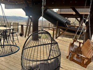 A wooden deck with a hanging chair, a swing, and a table with chairs. - Two Treehouses + Bridge | Double 20-Foot Slides | Smoky Mountain Views (Sevierville)