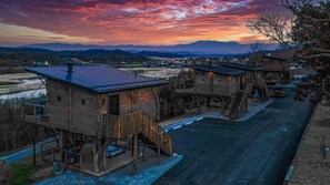 A series of wooden cabins with external staircases, connected by a paved walkway, set against a mountainous backdrop. - Two Treehouses + Bridge | Double 20-Foot Slides | Smoky Mountain Views (Sevierville)