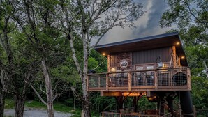 A two-story wooden cabin with a balcony, surrounded by trees. - Smoky Mountain Luxury Treehouse | 20-Foot Slide | Secret Beds | Sleeps 6 (Sevierville)