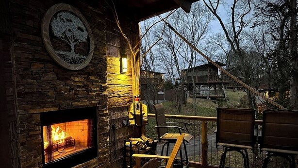 A cozy outdoor seating area with a stone fireplace, wooden deck, and chairs. - Smoky Mountain Luxury Treehouse | 20-Foot Slide | Secret Beds | Sleeps 6 (Sevierville)