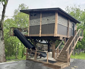 A wooden treehouse with a staircase and a slide, surrounded by trees.