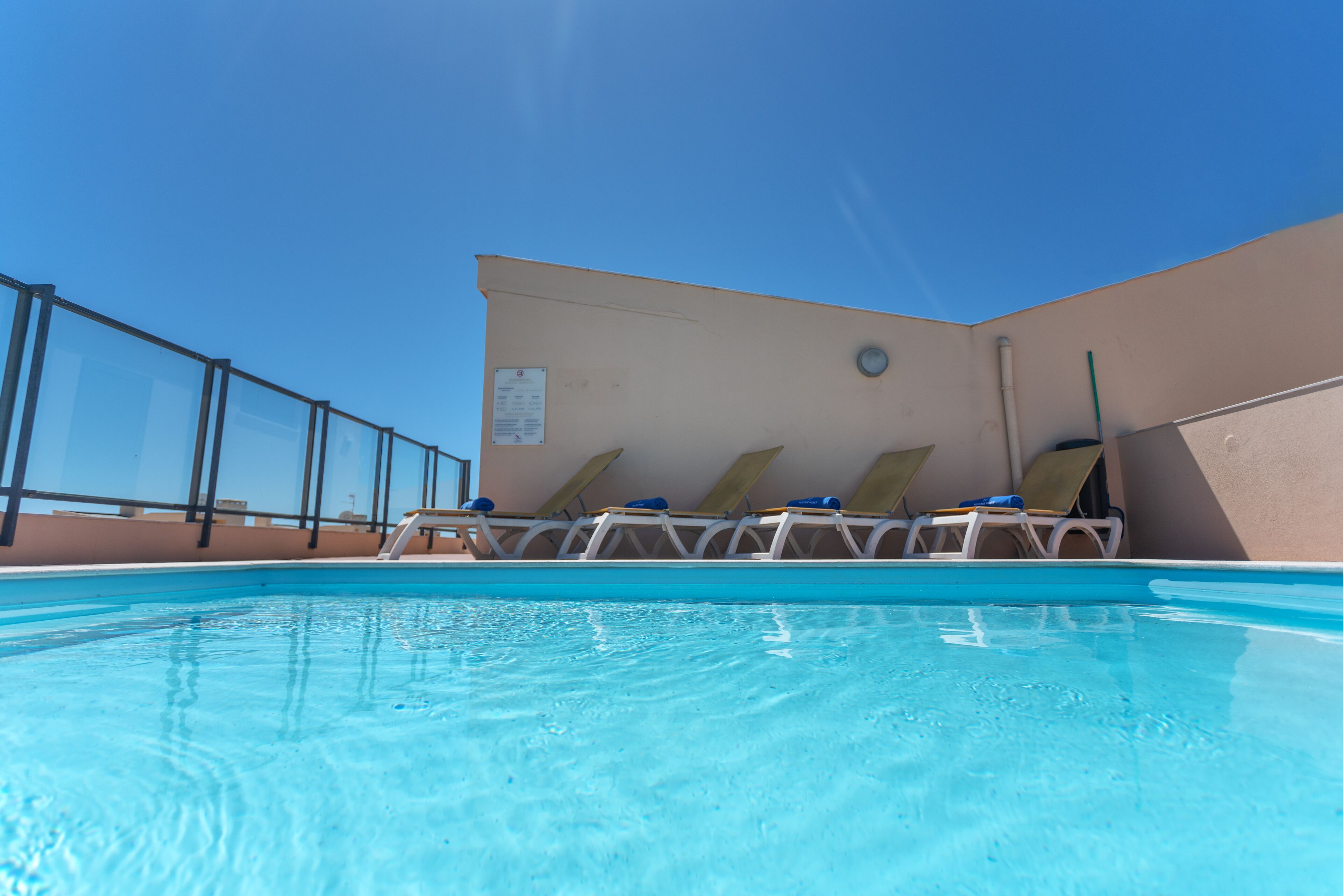 Una piscina con agua azul cristalina, rodeada de sillas de descanso y una barandilla de vidrio.