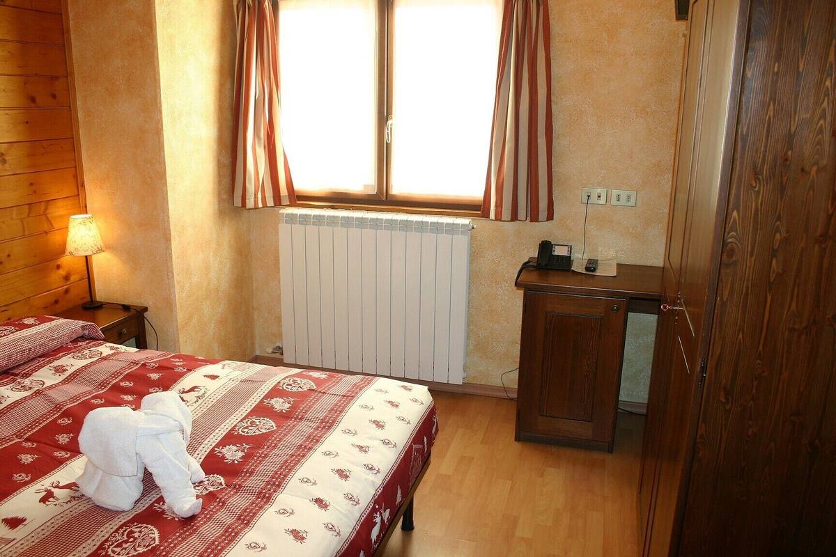 A bedroom with a wooden headboard, a bed with a patterned bedspread, a radiator, a wooden nightstand with a lamp, and a wooden cabinet with a telephone.